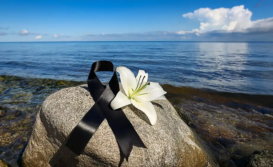 Schwarzes Trauerband und weiße Lilienblüten auf einem Felsen am ruhigen Meer